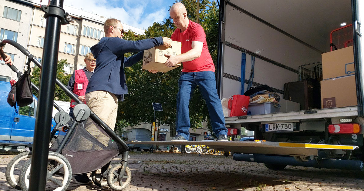 Mies nostaa ruskeaa pahvilaatikkoa lastenrattaista kuorma-autoon, toinen mies auttaa häntä.