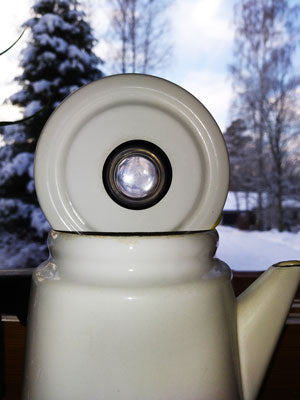 Valkoinen kahvipannu ikkunalaudalla sivultapäin, kannen nuppi on läpinäkyvä, taustalla talvimaisemaa.