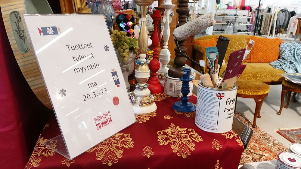 Maalisiveltimiä pystyssä maalipurkissa pöydällä, taustalla myymälää ja keltainen vintage-sohva.