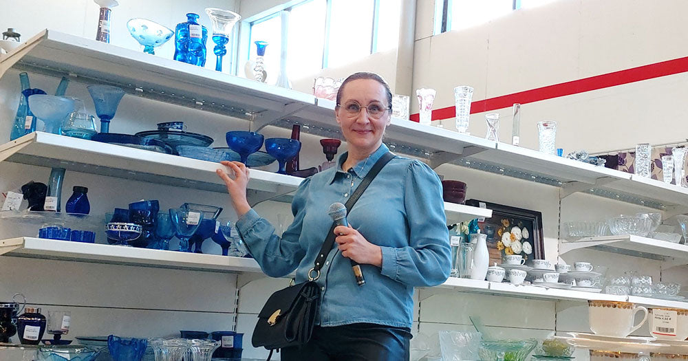 Siniasuinen henkilö sinisten lasiesineiden hyllyn edessä myymälässä, mikrofoni kädessään