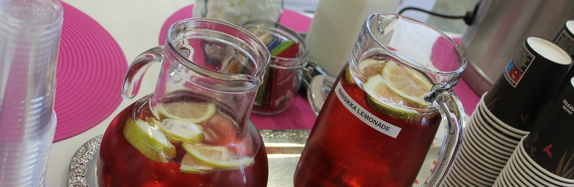 Tarjoilupöydällä mehukannuissa punaista juomaa ja sitruunaviipaleita, toisessa kannussa teksti "mansikka lemonade". Taustalla kahvipöydän tarjoiluja: teepusseja, palasokereita, pahvikuppeja. 