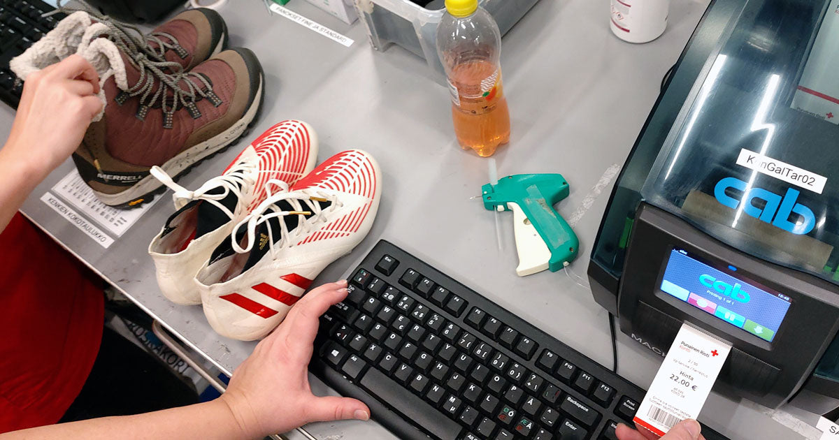 Jalkapallonappikset ja lasten talvikengät hinnoittelukoneen edessä. Koneesta tulostuu ulos lappu, jossa hinta 22 €.