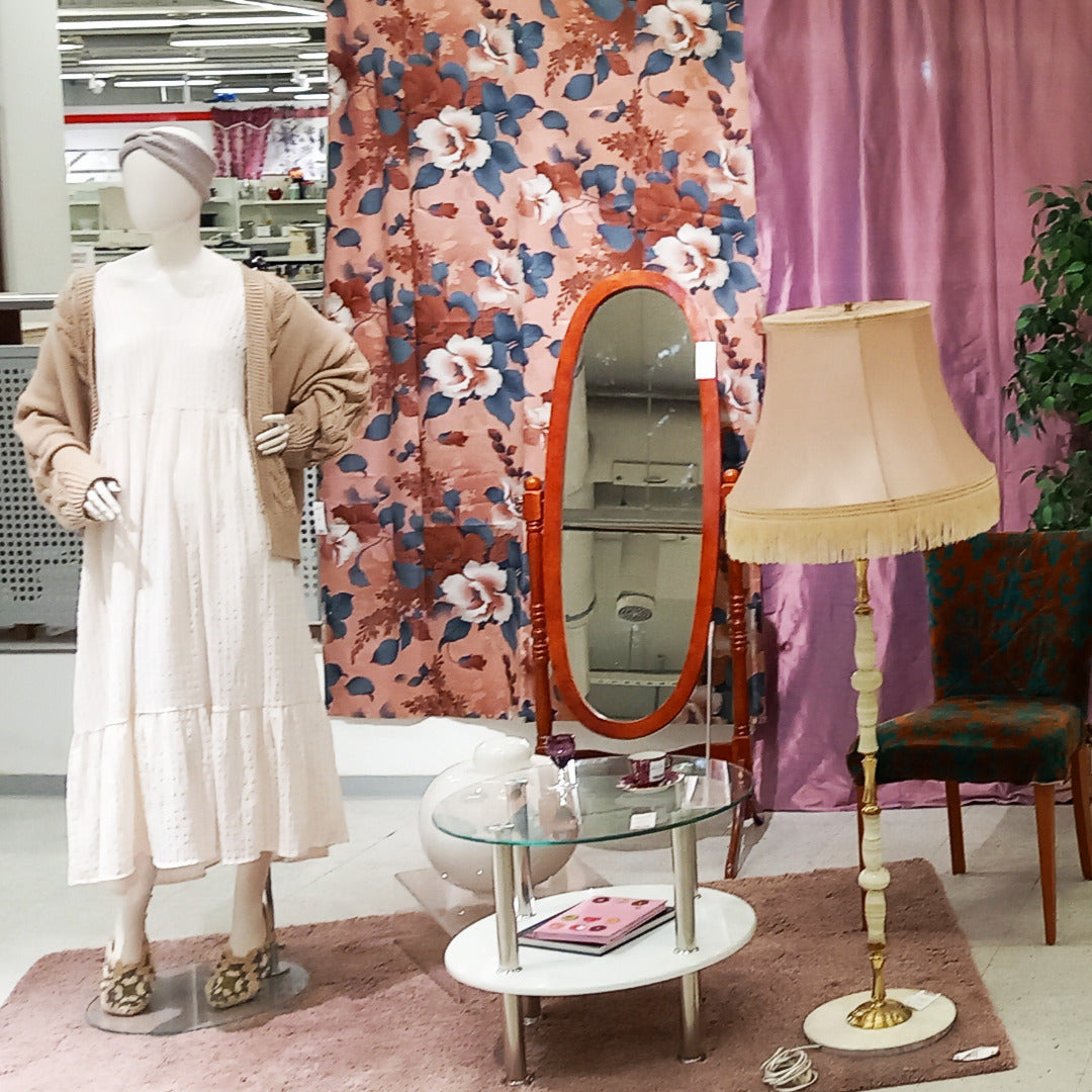 Retrohenkinen stailaus, jossa on vanha jalkalamppu, kaksitasoinen pikkupöytä ja jalallinen vartalopeili, taustalla kukkaverho. Mallinukella on mekko ja villatakki.