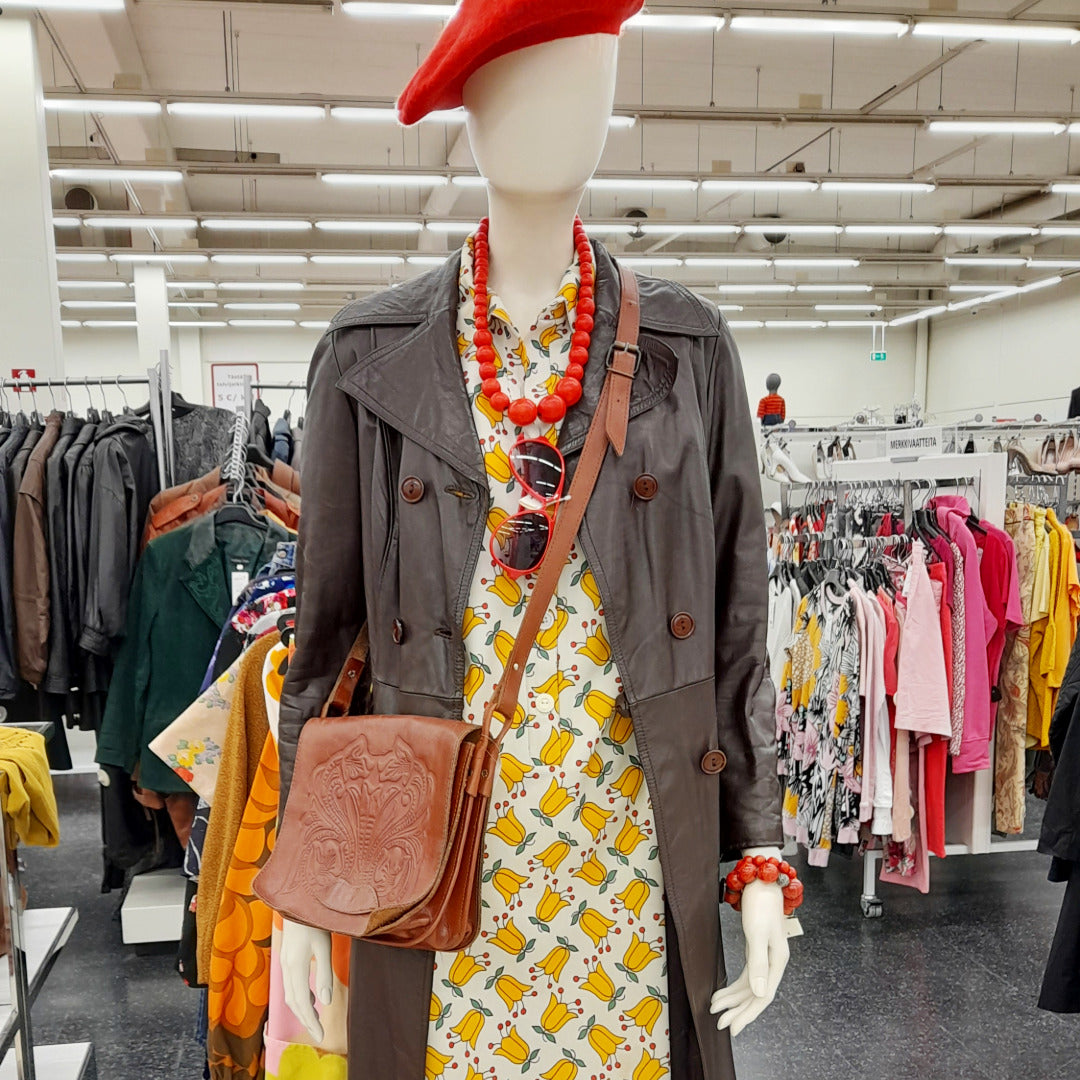 Mallinukella trenssitakki, nahkainen olkalaukku ja punainen baskeri ja helmet. Taustalla näkymä vaateosastolle.